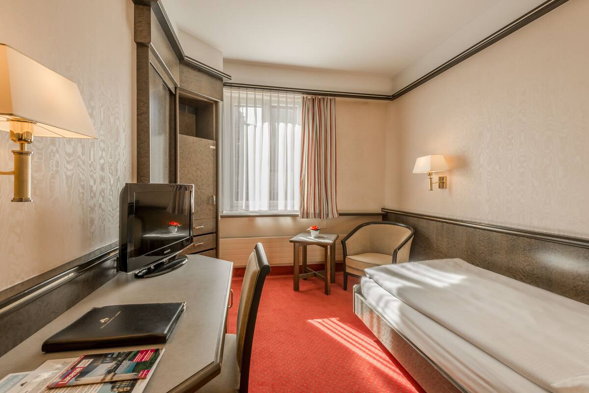 Hotellihuone, jossa on sänky, työpöytä kannettavalla tietokoneella, tuoli, televisio ja ikkuna, jossa on verhot.