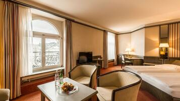 A hotel room with a bed, a desk with a television, a dining table with fruit, and a window with curtains.