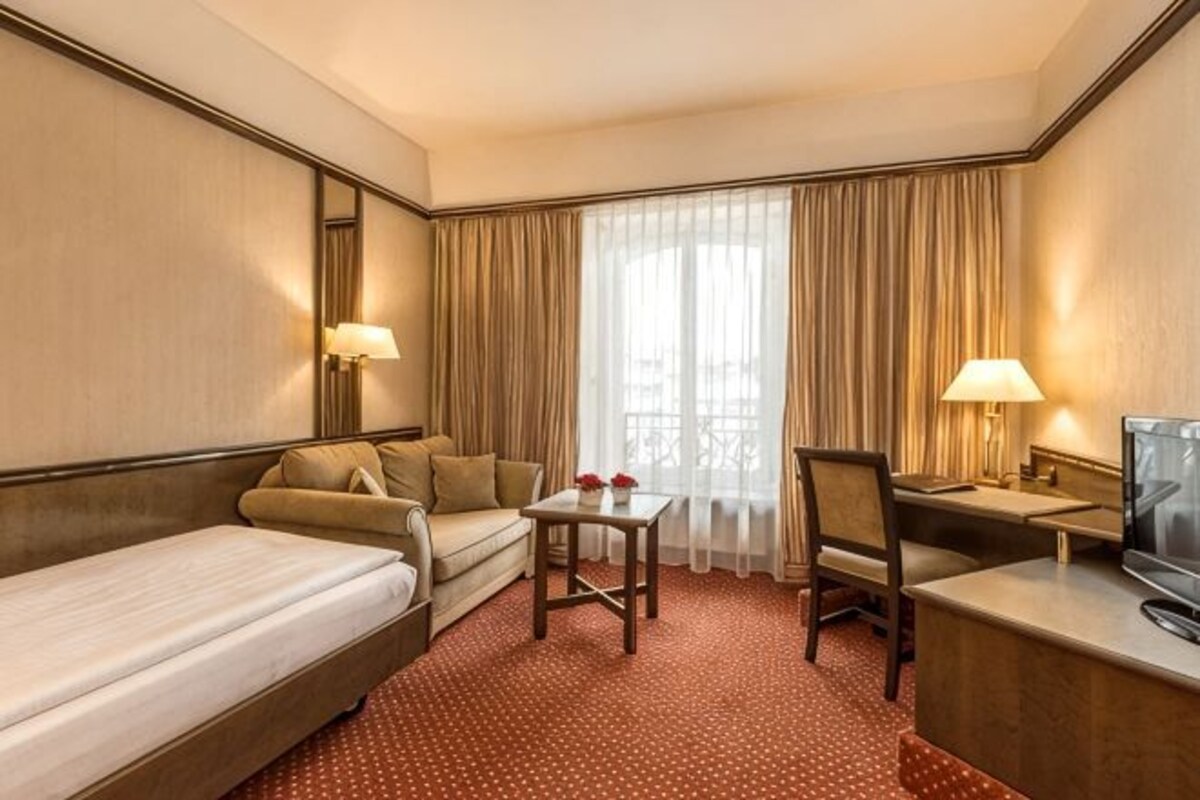 A hotel room with a bed, a sofa, a small table, a chair, a desk, a television, and curtains.