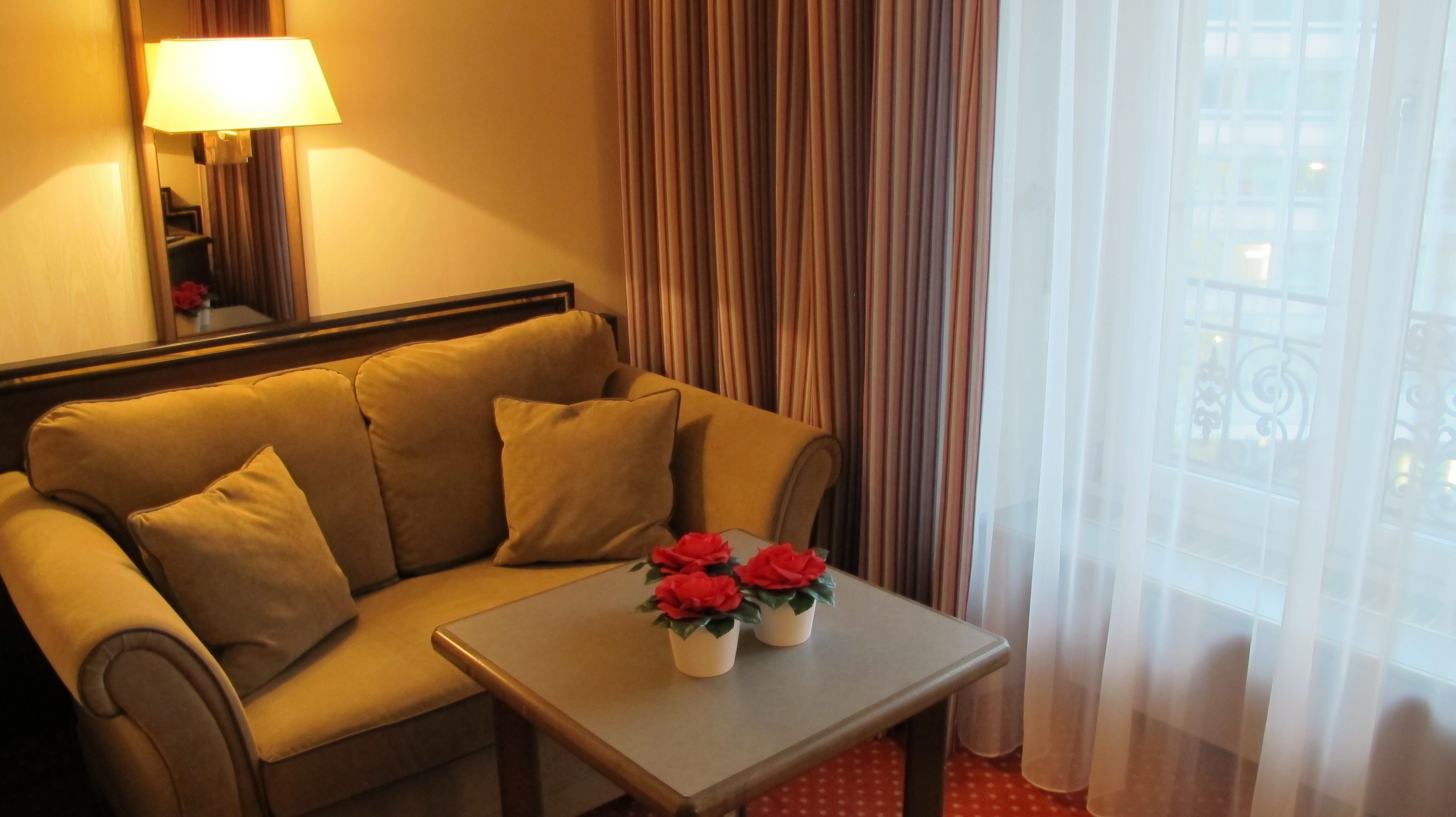 Habitación de hotel con sofá, una mesa con dos macetas, una lámpara y vistas a través de cortinas de gasa.