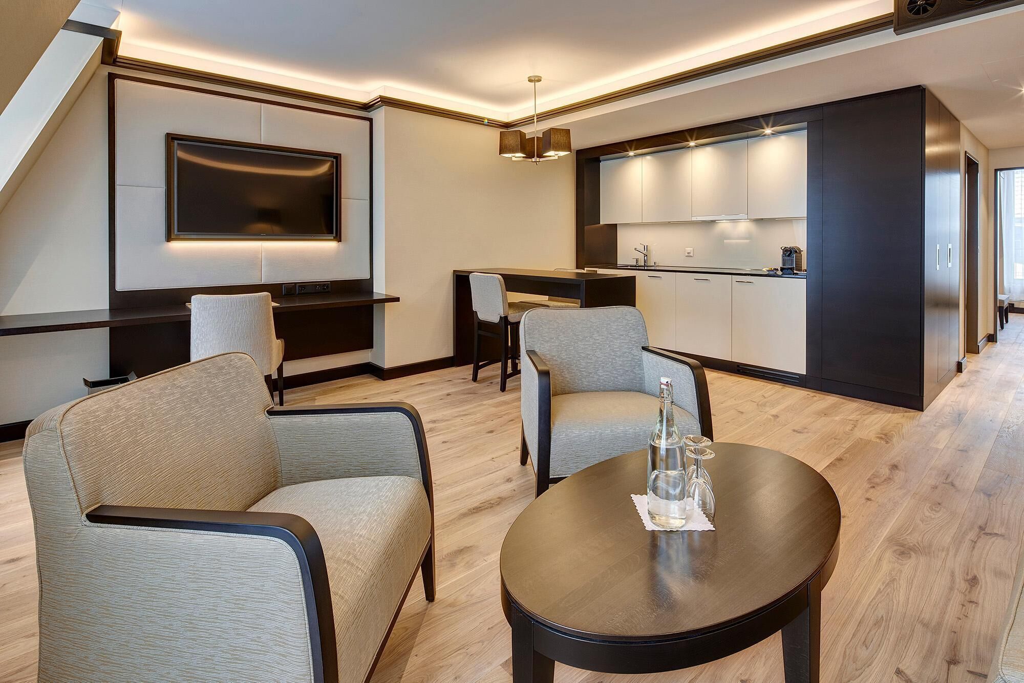 A modern living room with a flat-screen TV, a dining area, and a kitchen with dark cabinets.