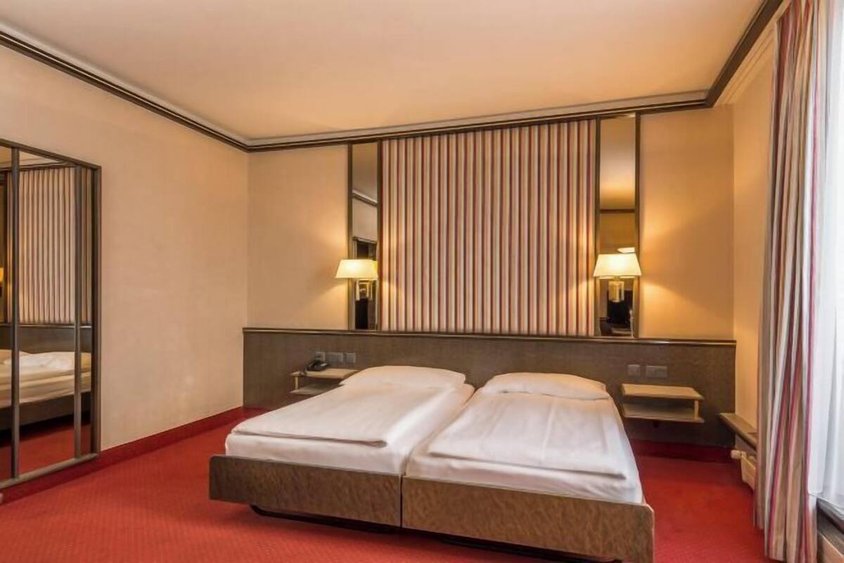 Hotellihuone, jossa on parivuode, yöpöytä, peili ja punaisella matolla päällystetty lattia.
