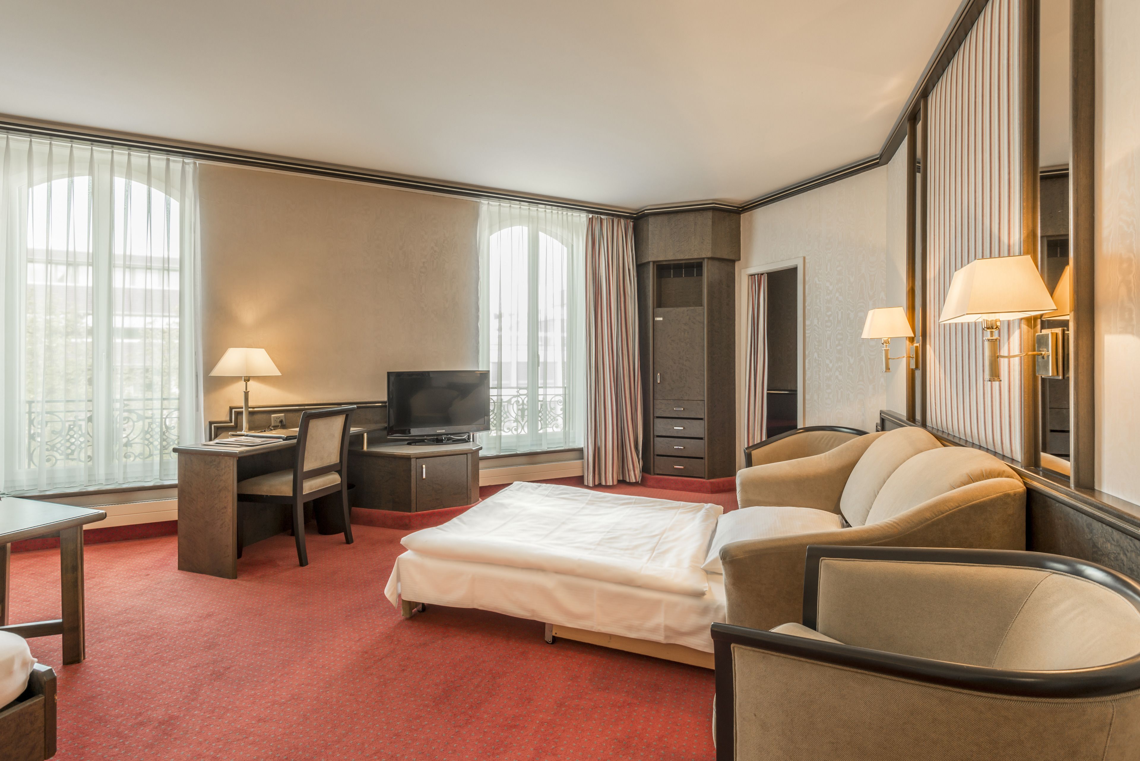 Habitación de hotel con cama, escritorio, silla, televisor y ventana grande con cortinas.