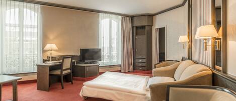 Habitación de hotel con cama, escritorio, silla, televisor y ventana grande con cortinas.