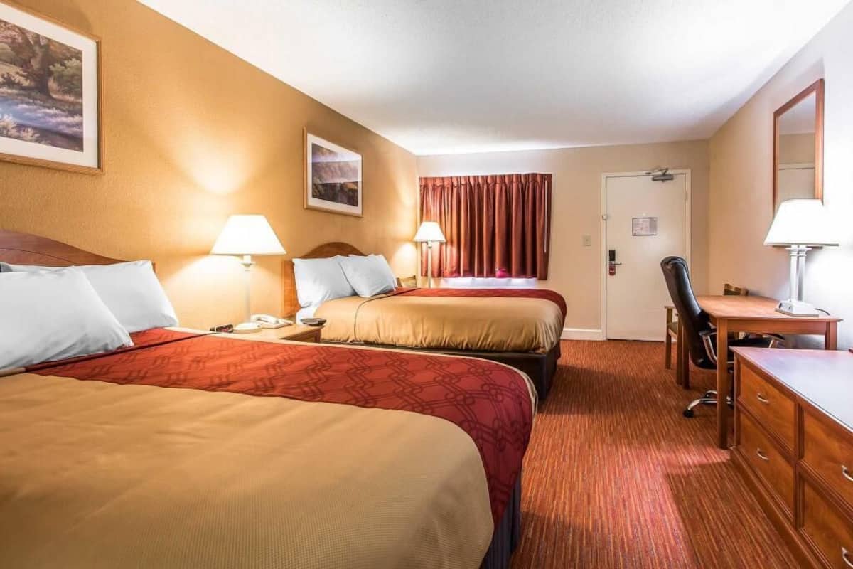A hotel room with two beds, a desk, a chair, a lamp, and a door.