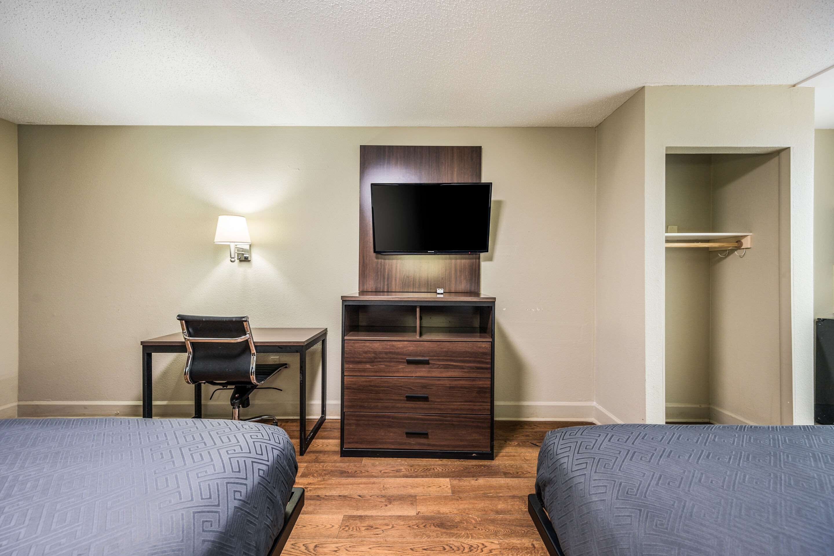 A hotel room with two beds, a desk, a chair, a TV mounted on a wooden cabinet, and a wall-mounted lamp.