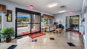 Un vestíbulo de hotel con un amplio ventanal que da al exterior, un sofá, una mesita, una planta en maceta y una alfombra roja con el nombre del hotel.