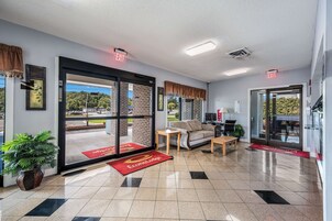 A hotel lobby with a large glass door leading outside, a sofa, a small table, a potted plant, and a red carpet with the hotel name.