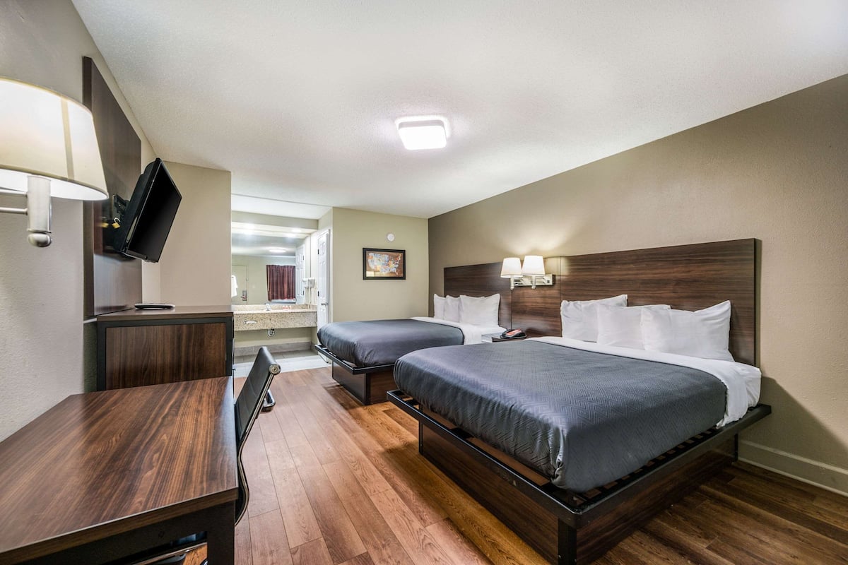 A hotel room with two beds, a desk, a TV, and a bathroom visible in the background.