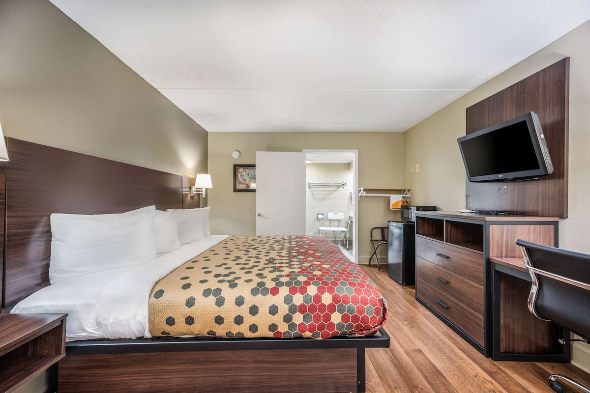 A hotel room with a bed, a desk, a chair, a television, and a bathroom visible through an open door.