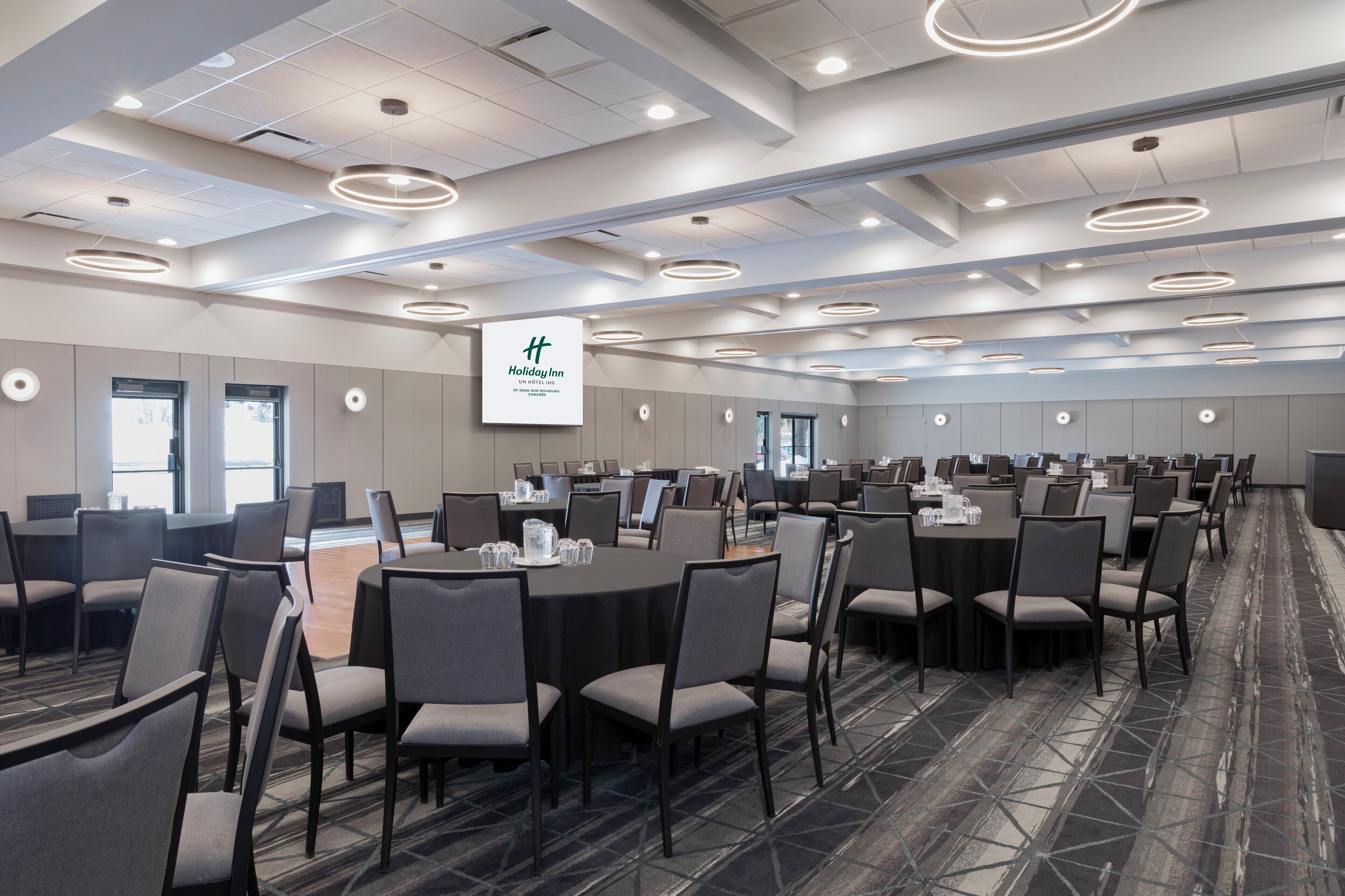 Ein großer Konferenzraum mit mehreren Tischen und Stühlen, einer abgehängten Deckenbeleuchtung und einem großen Bildschirm, der das Holiday Inn Logo zeigt.