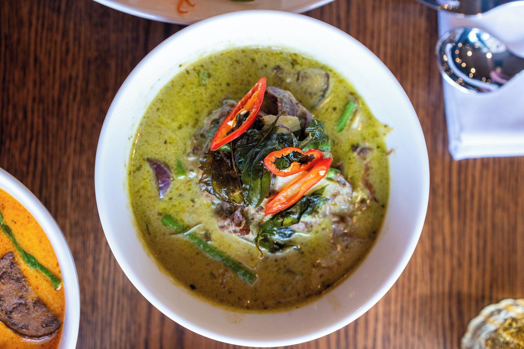 A bowl of green curry with red chili slices and leafy greens, served on a wooden table.
