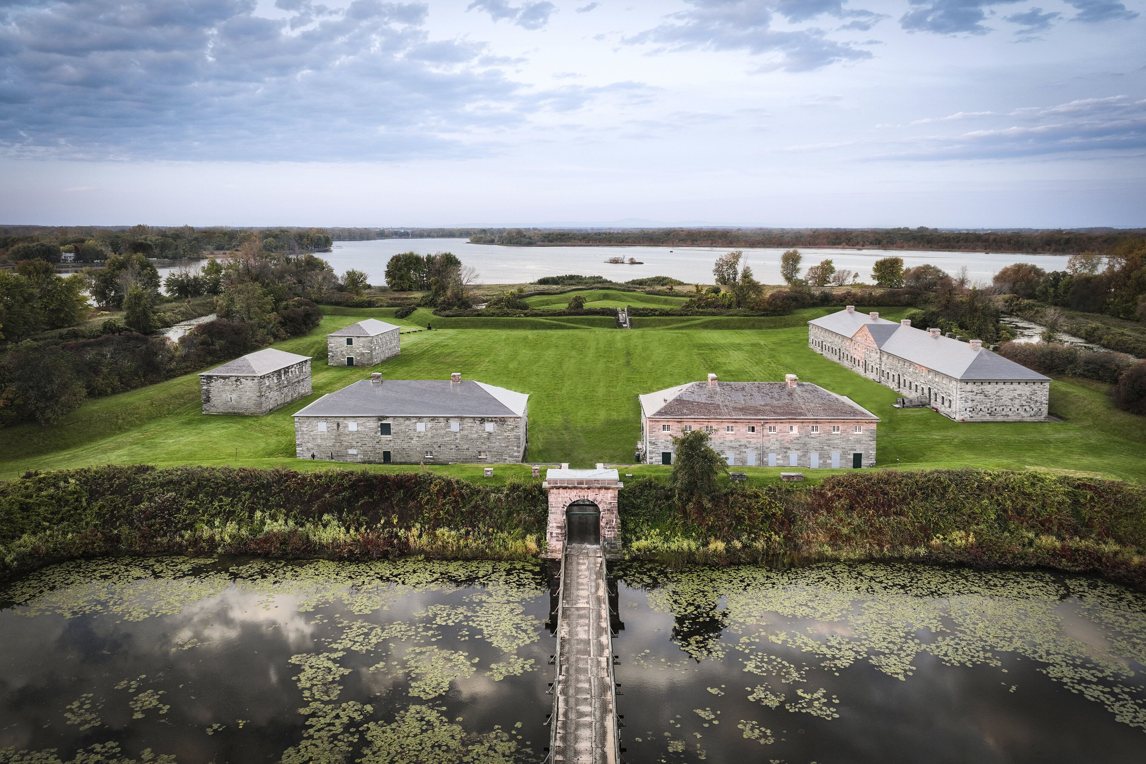 A large estate with multiple stone buildings, a central lawn, and a body of water with a bridge leading to the buildings.