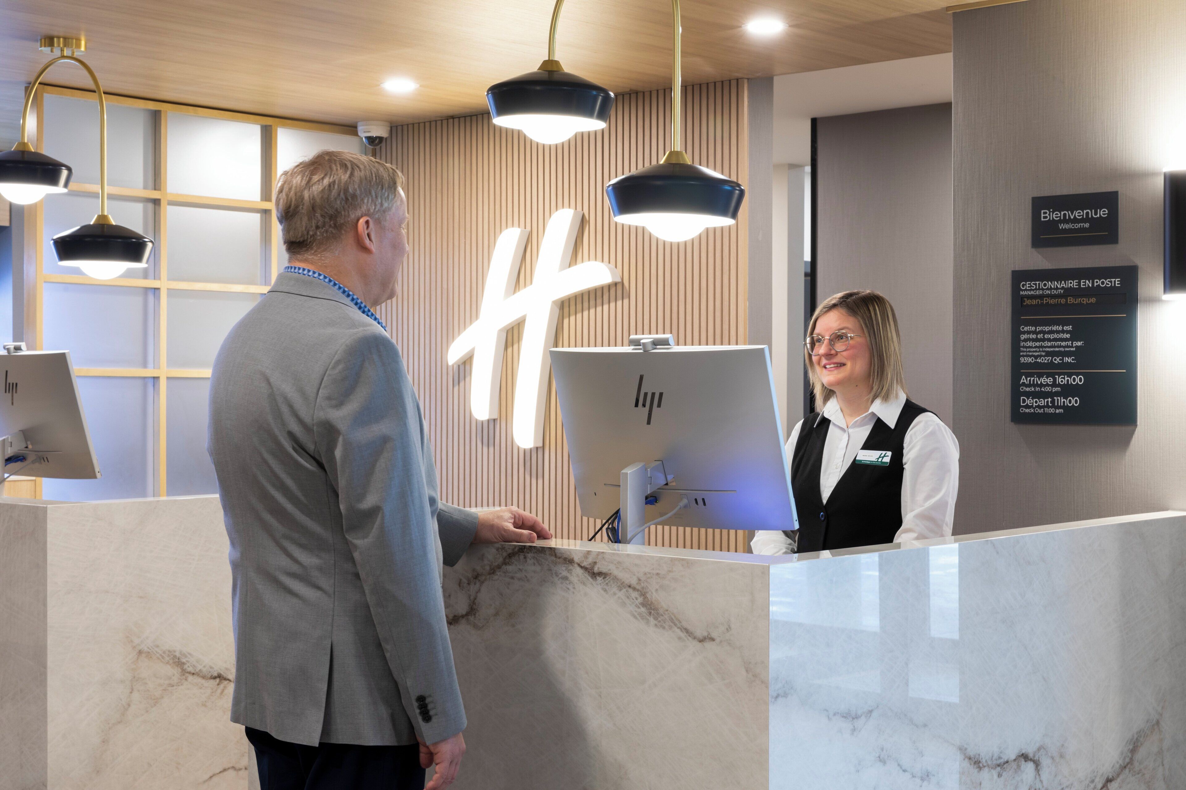 Un comptoir d’accueil avec un plateau en marbre, un grand panneau lumineux en forme de « H » et un écran numérique affichant les heures d’arrivée et de départ.