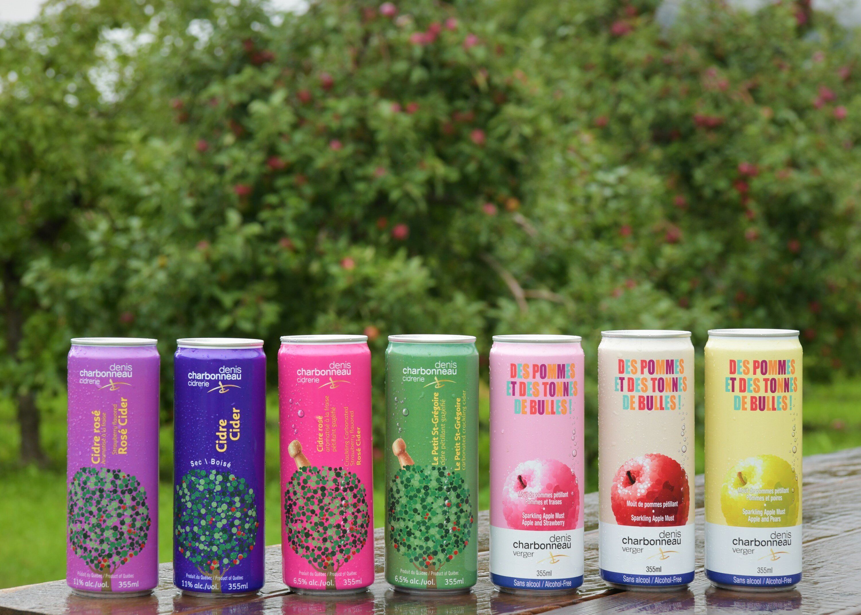 Seven cans of sparkling apple cider in various colors, each with a different label, placed on a wooden surface outdoors with a blurred green background.