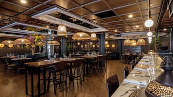 A restaurant with wooden tables and chairs, hanging wicker lamps, and a marble bar counter.
