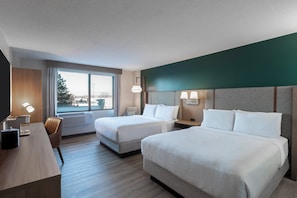 Two single beds with white bedding, a desk, and a chair in a stylish room with a city view.