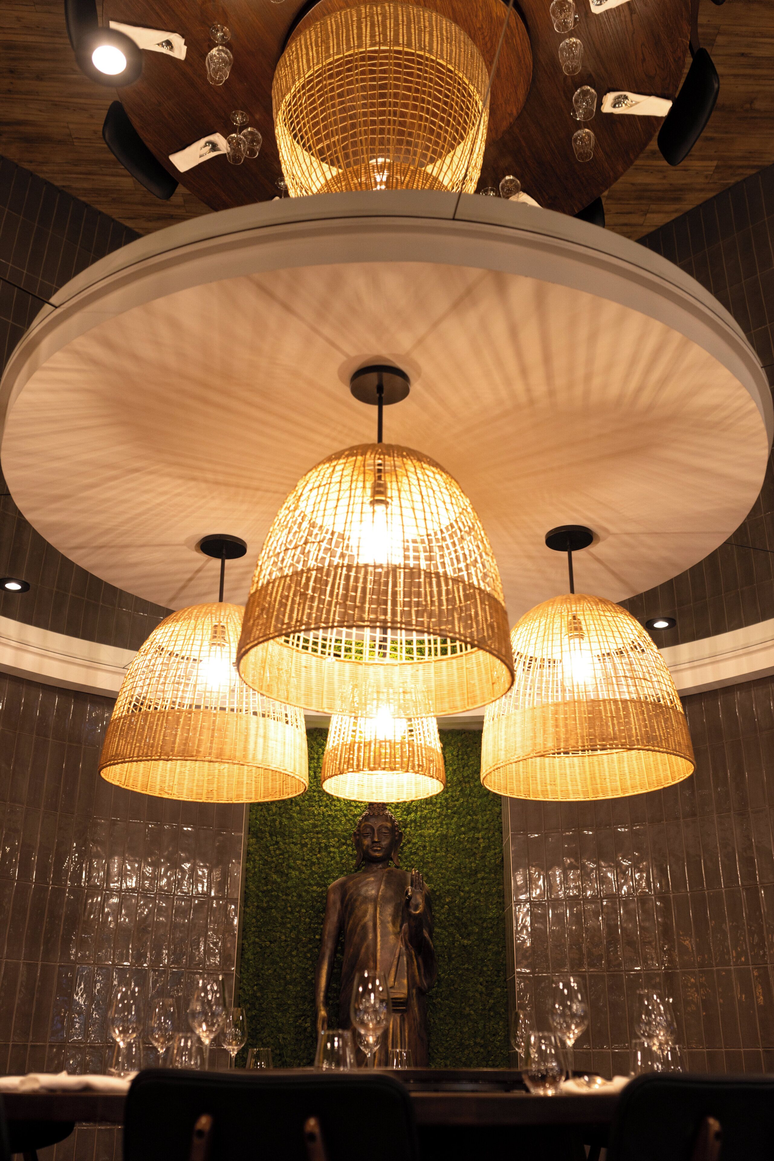 A restaurant interior with a Buddha statue, round tables, and hanging wicker lamps.