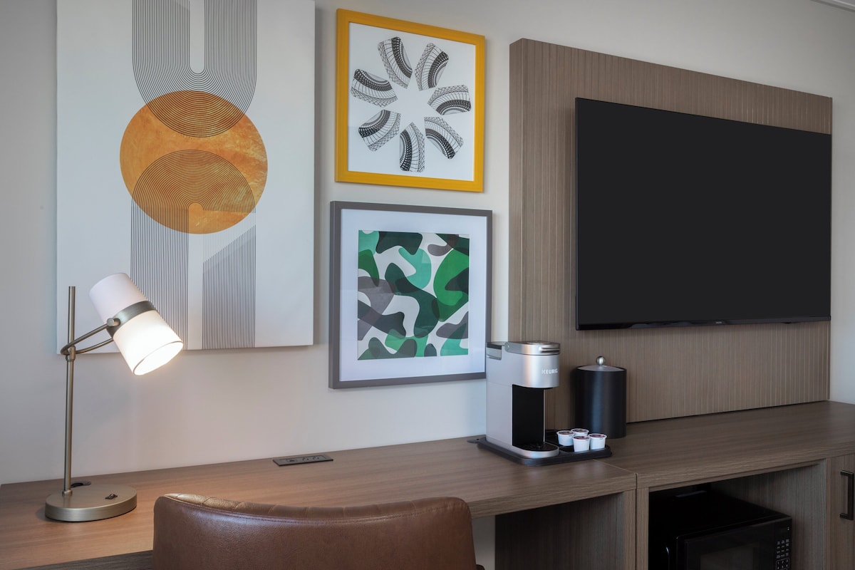 A hotel room with a wooden desk, a Keurig coffee maker, a flat-screen TV, and three framed artworks on the wall.