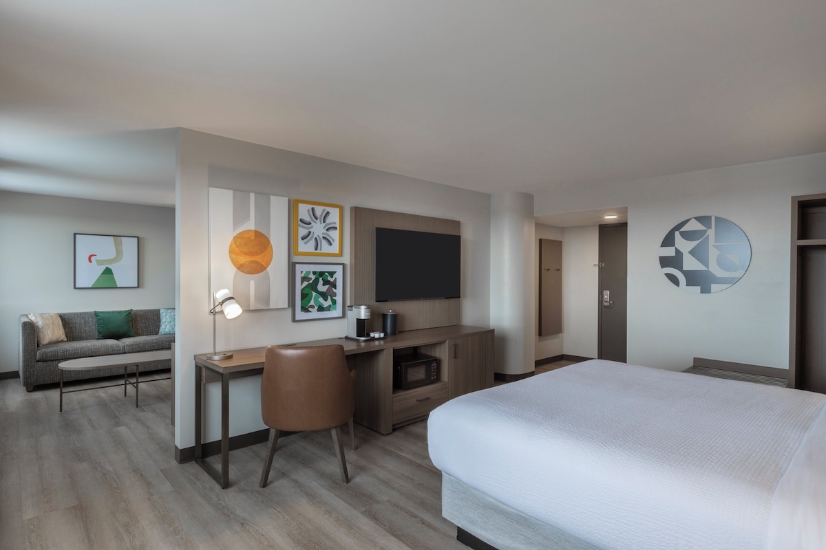 A hotel room with a bed, a desk with a chair, a TV, and artwork on the walls.