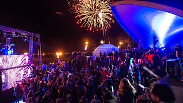 Evento al aire libre concurrido, con escenario, pantalla grande y fuegos artificiales al fondo.