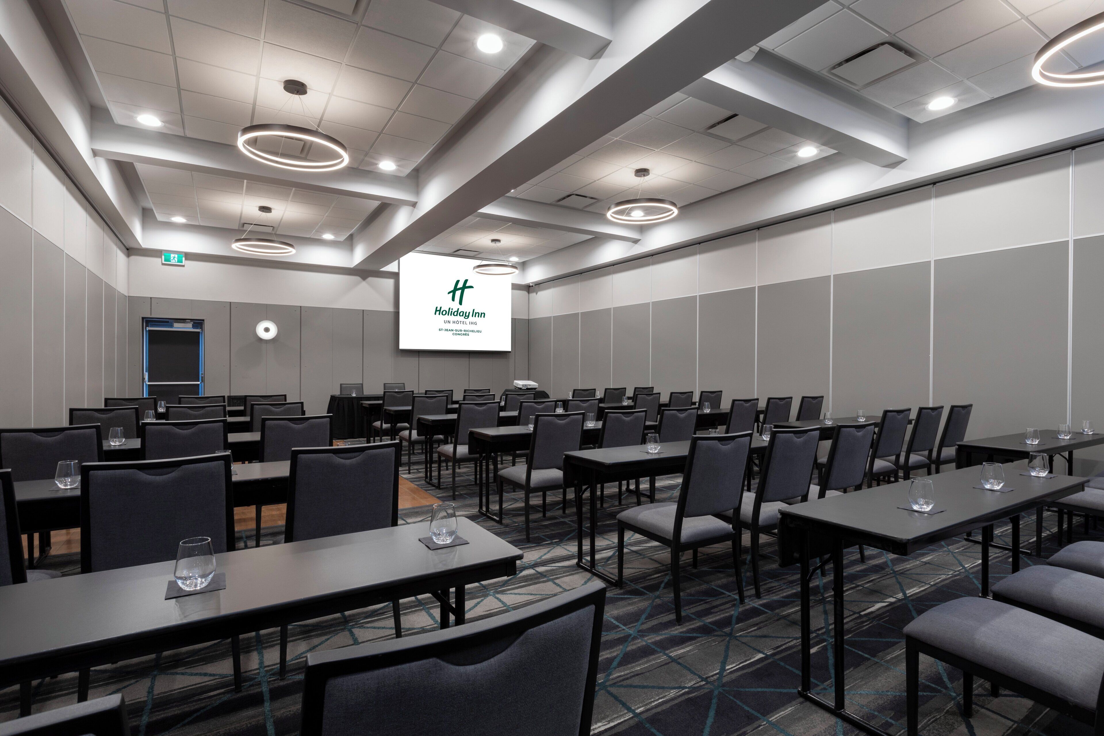 A conference room with multiple tables and chairs, a large screen displaying a logo, and a modern ceiling design.