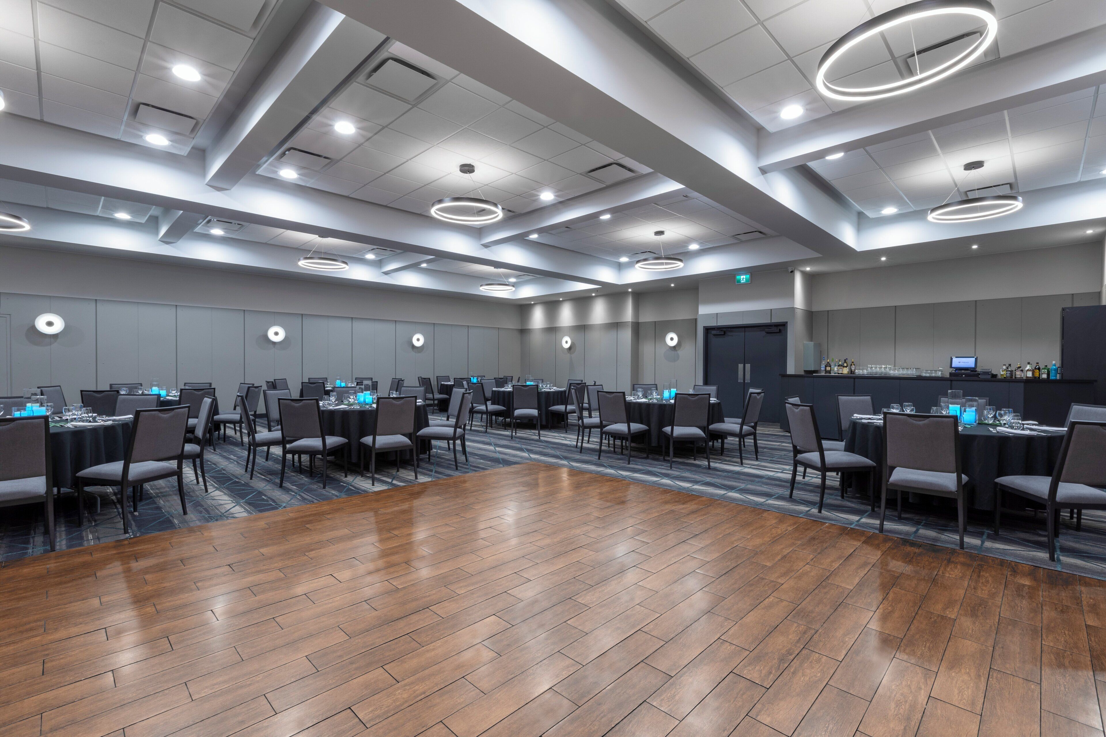 Une salle de conférence moderne avec un plancher en bois, des murs gris et des luminaires circulaires au plafond. Des tables sont disposées, accompagnées de chaises et d’ordinateurs portables.