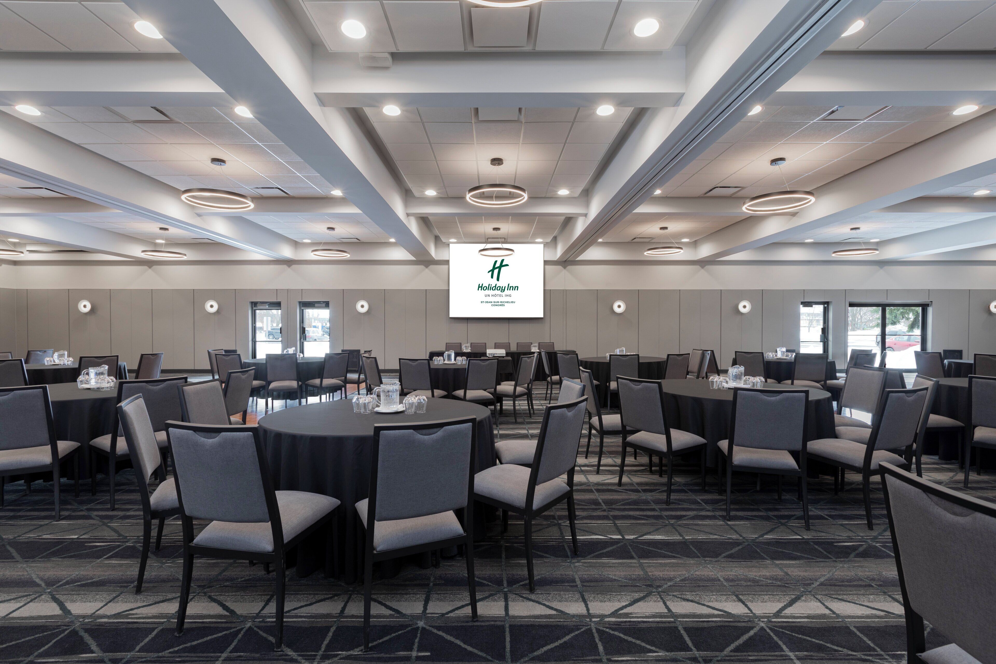 Une salle de conférence dotée de tables et de chaises rondes, d’un grand écran affichant un logo et d’un design de plafond moderne.