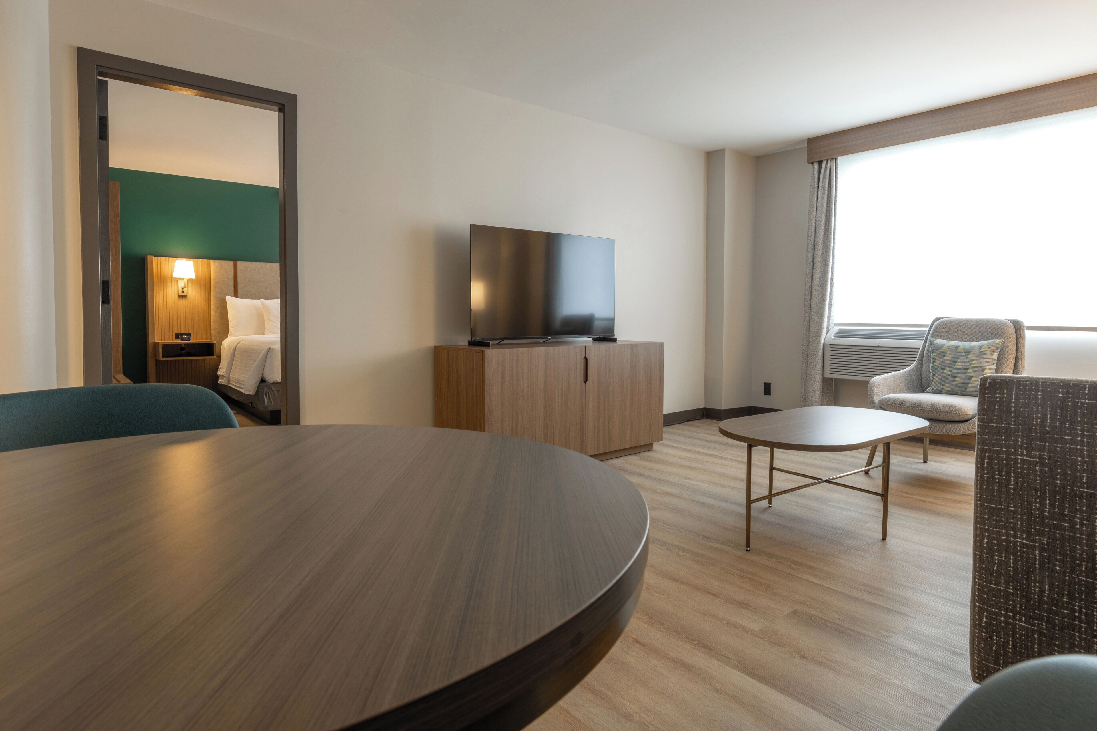 Una habitación de hotel moderna con una mesa redonda para comer, un televisor de pantalla plana sobre un mueble de madera, un sofá y un ventanal grande.