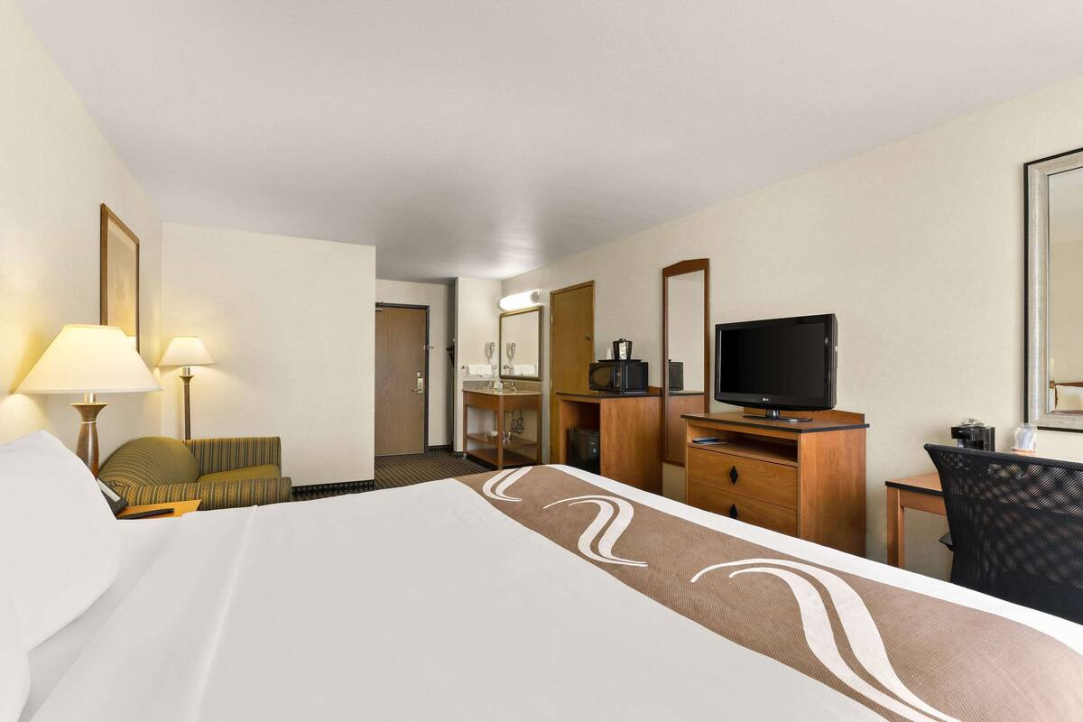 Ein Hotelzimmer mit einem großen Bett, einem Schreibtisch mit Computer, einem Fernseher und einem durchgegangenen Tür angedeuteten Badezimmer.