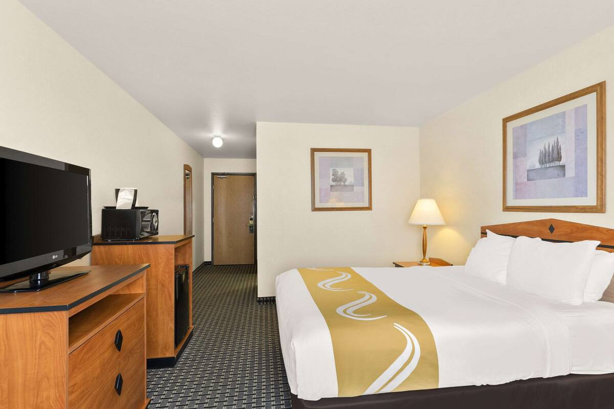 Ein Hotelzimmer mit einem Bett, einem Fernseher, einem Schreibtisch mit Lampe und gerahmten Bildern an den Wänden.