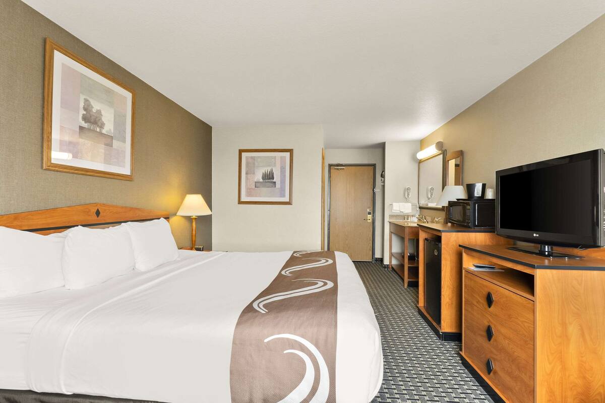 Ein Hotelzimmer mit einem großen Bett, einem Fernseher, einem Schreibtisch mit einer Mikrowelle und einem durch eine offene Tür einsehbaren Badezimmer.