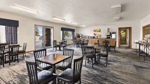A hotel dining area with multiple tables and chairs, a kitchen in the background, and large windows allowing natural light. - Quality Inn Cheyenne I-25 South (Cheyenne)