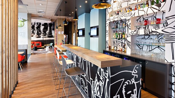 A modern bar area with a wooden counter, bar stools, and a wall with abstract black and white artwork. - ibis Agen Centre (Agen)