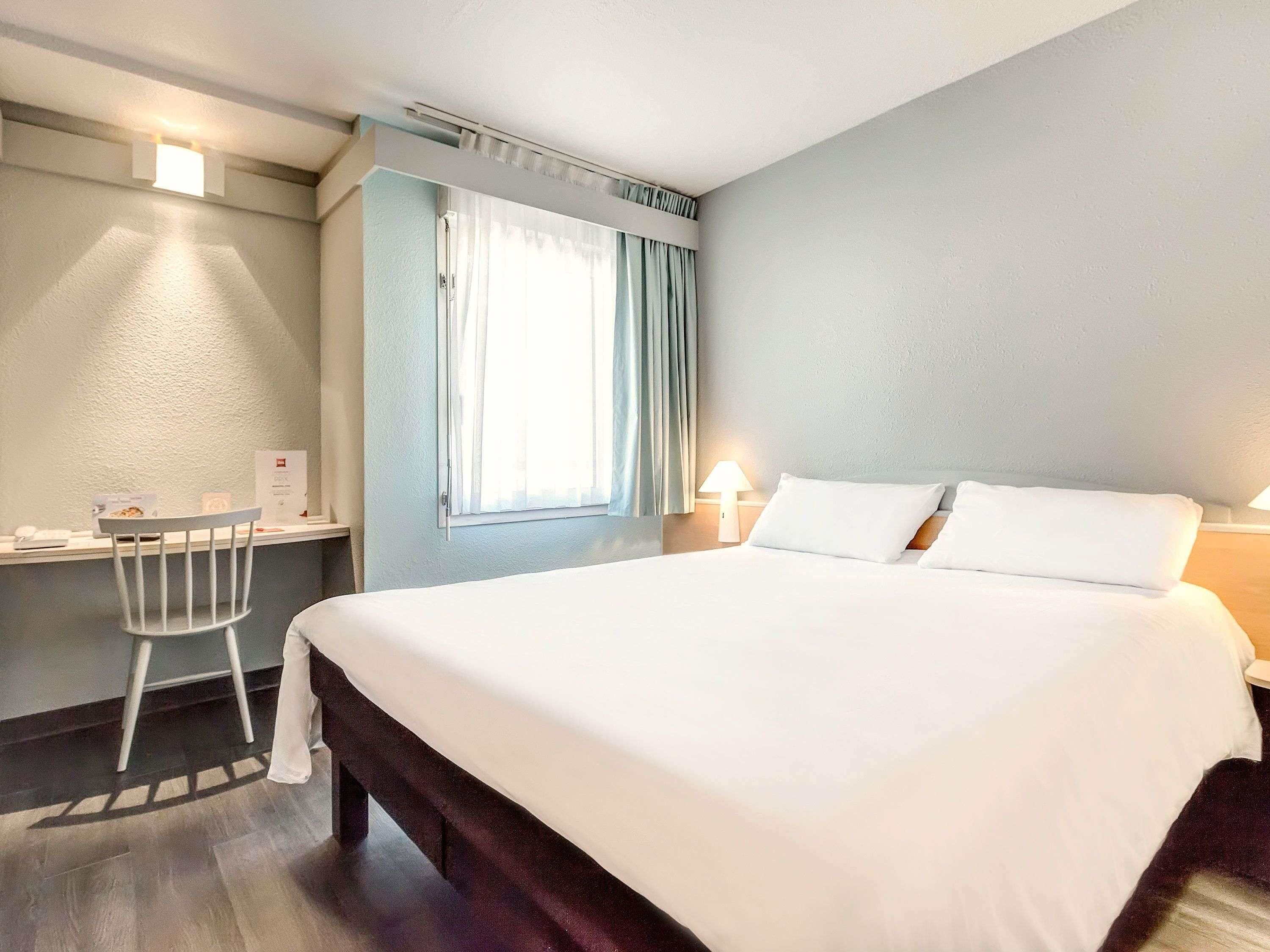 A hotel room with a bed, a desk, a chair, a window with curtains, and a wall-mounted lamp.