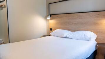 Una cama bien tendida con sábanas blancas, cabecera de madera y lámpara de noche.