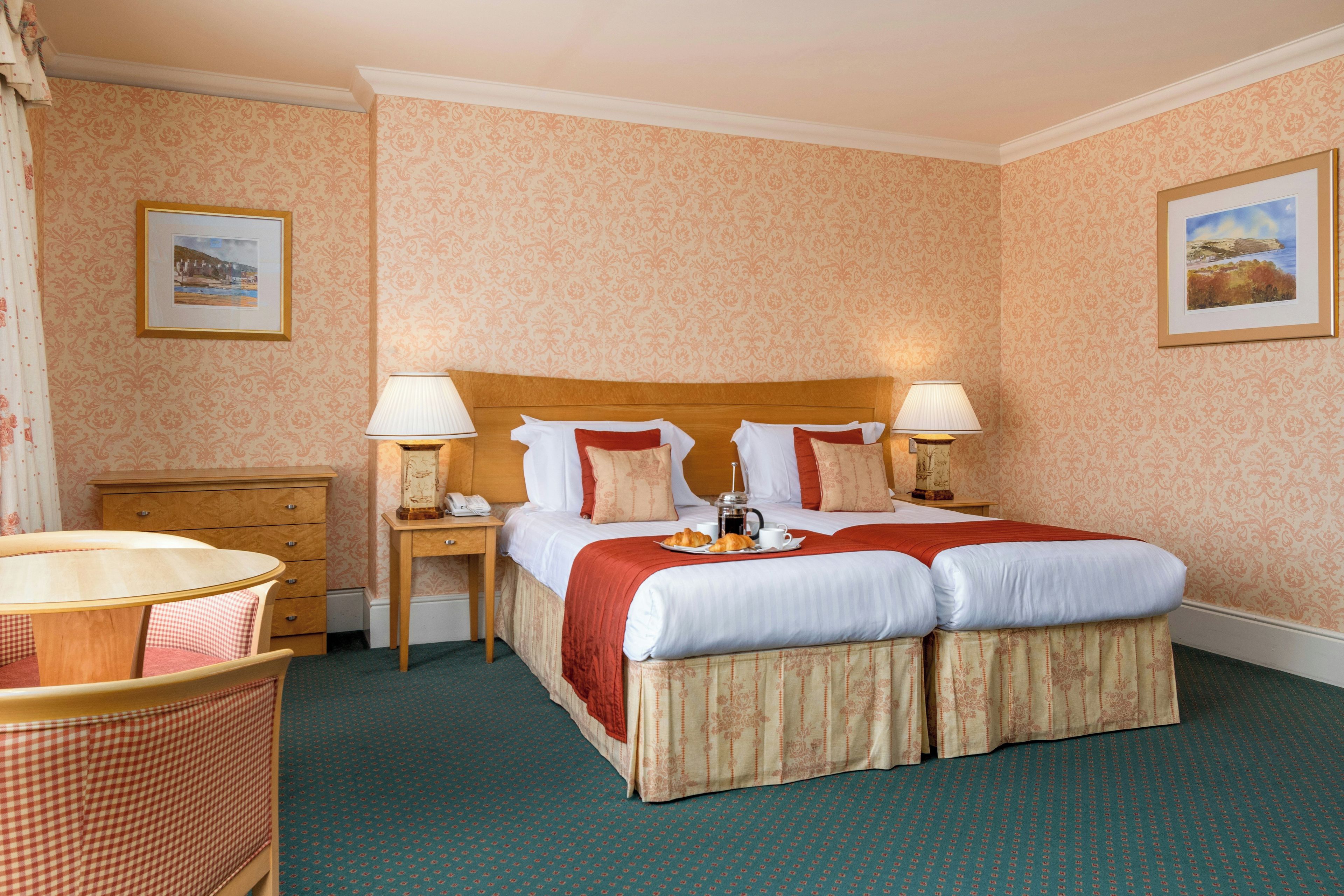 A hotel room with a double bed, two bedside lamps, a wooden nightstand, a small table with a lamp, a chair, and a checkered bench.