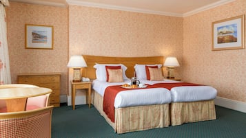 A hotel room with a double bed, two bedside lamps, a wooden nightstand, a small table with a lamp, a chair, and a checkered bench.