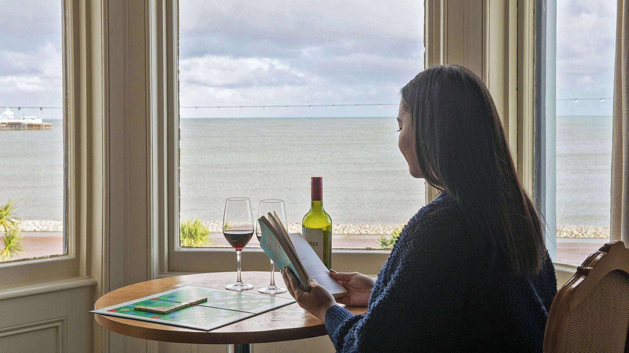 A person reading a book at a table by a window with a view of the sea.