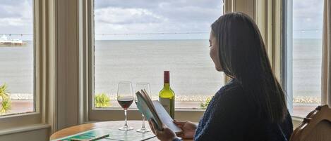 A person reading a book at a table by a window with a view of the sea.