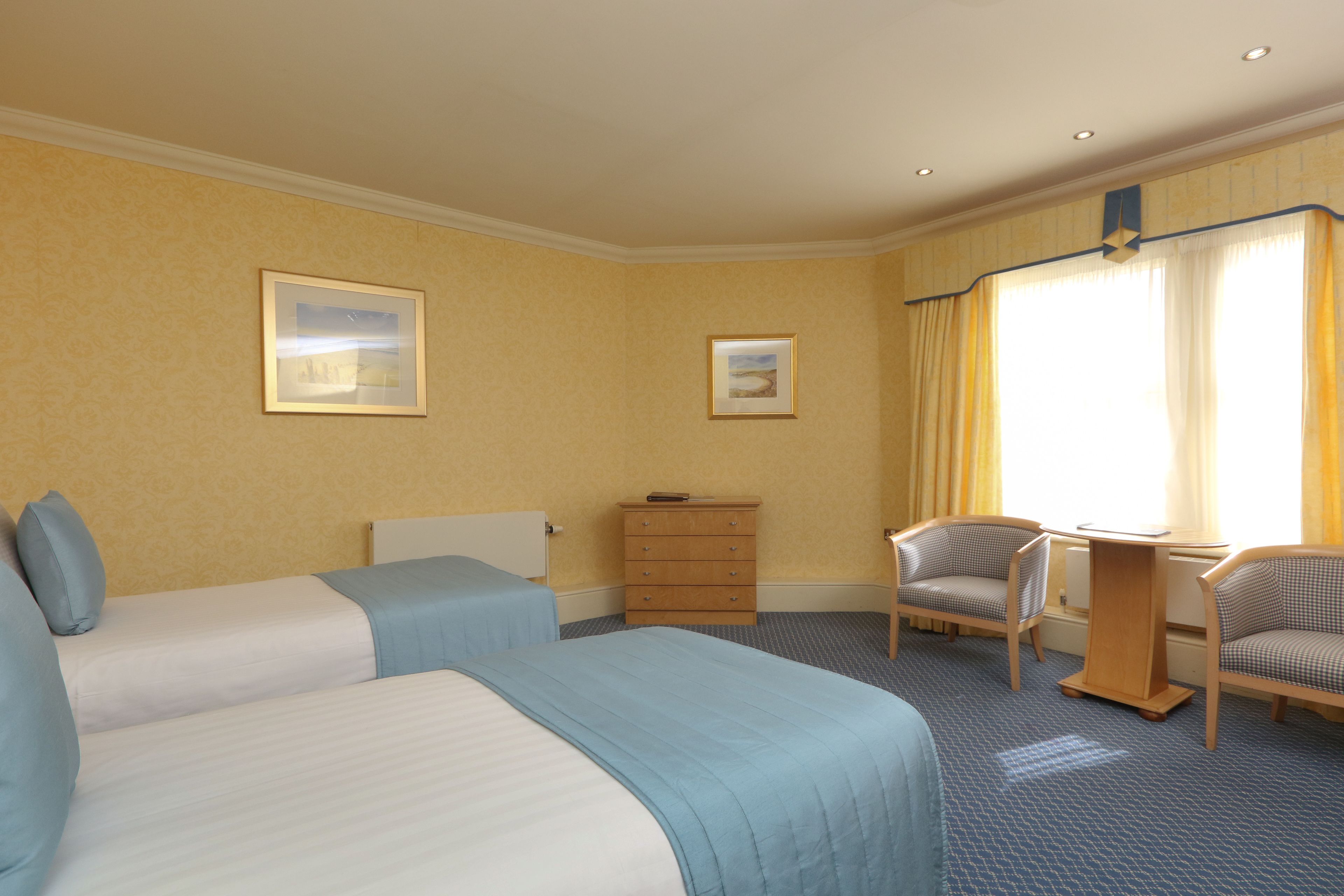 A hotel room with two beds, a wooden dresser, two armchairs, and two framed pictures on the wall.
