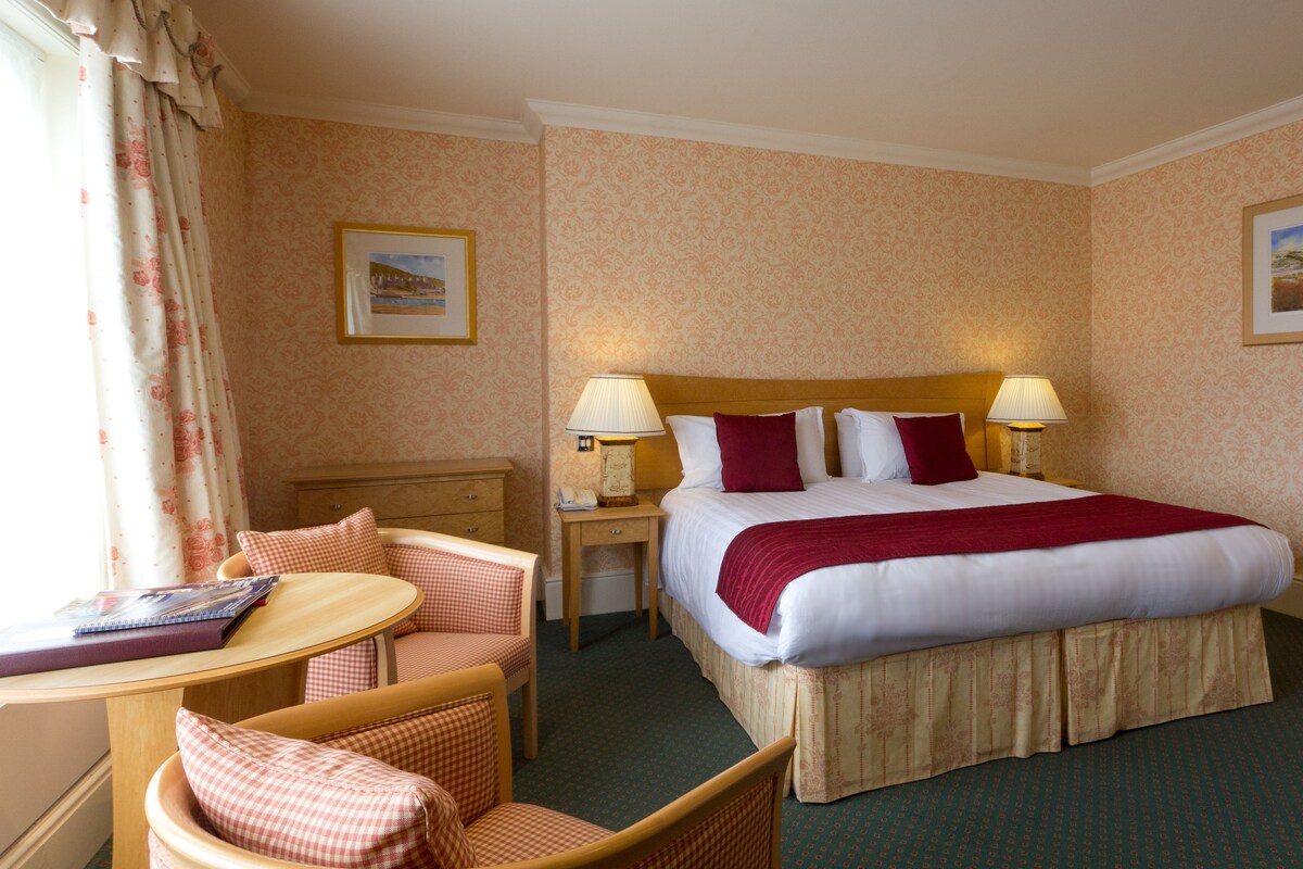 A hotel room with a large bed, two bedside lamps, a wooden nightstand, a chair, a small table, and two framed pictures on the walls.