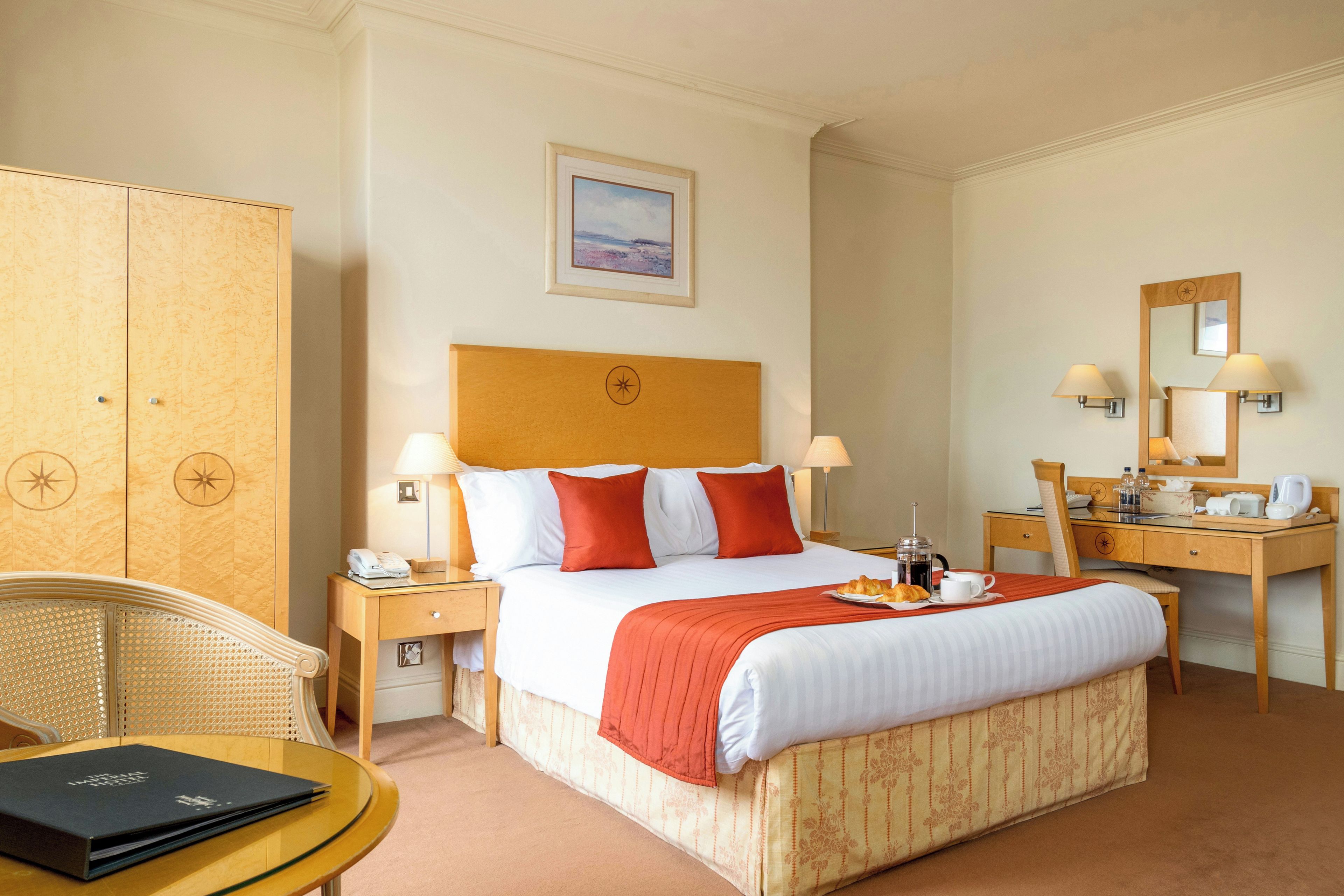 A hotel room with a large bed, a desk, a chair, a wardrobe, and a small table with a book.