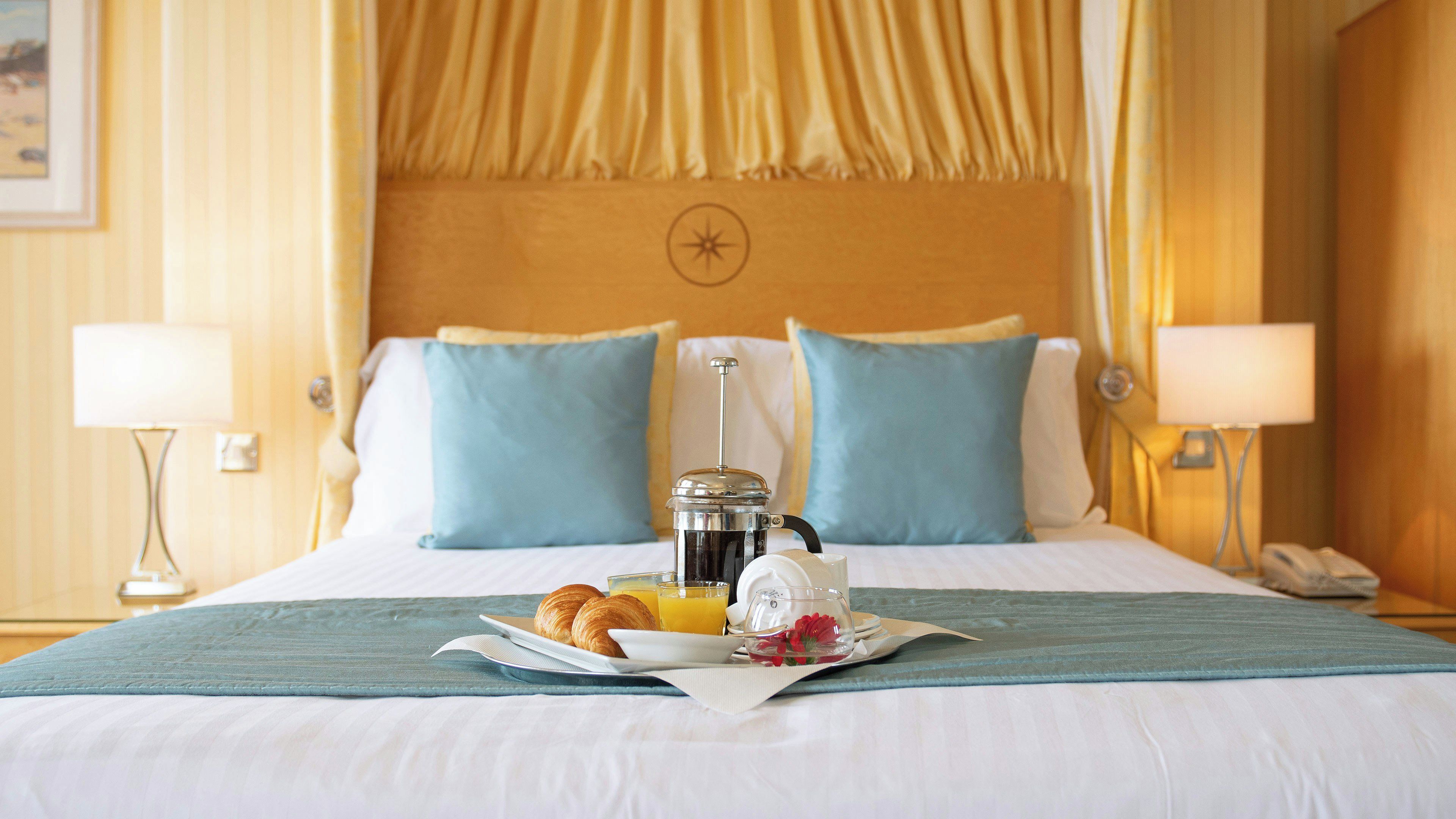 A hotel room with a bed, a coffee maker, croissants, and a glass of orange juice on the bed.