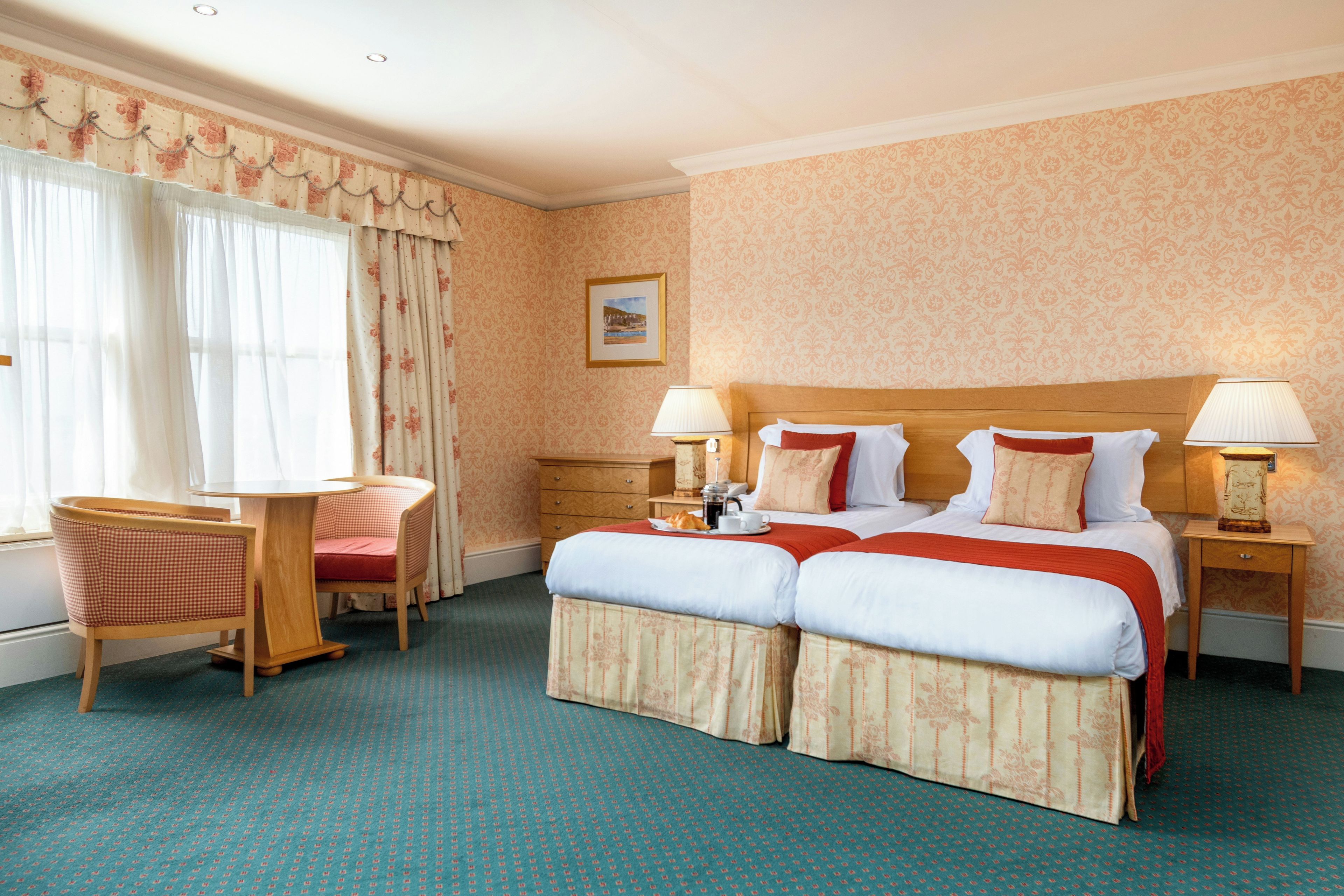 A hotel room with a double bed, two bedside tables with lamps, a small wooden chair, a table with a tray of breakfast items, and a window with curtains.