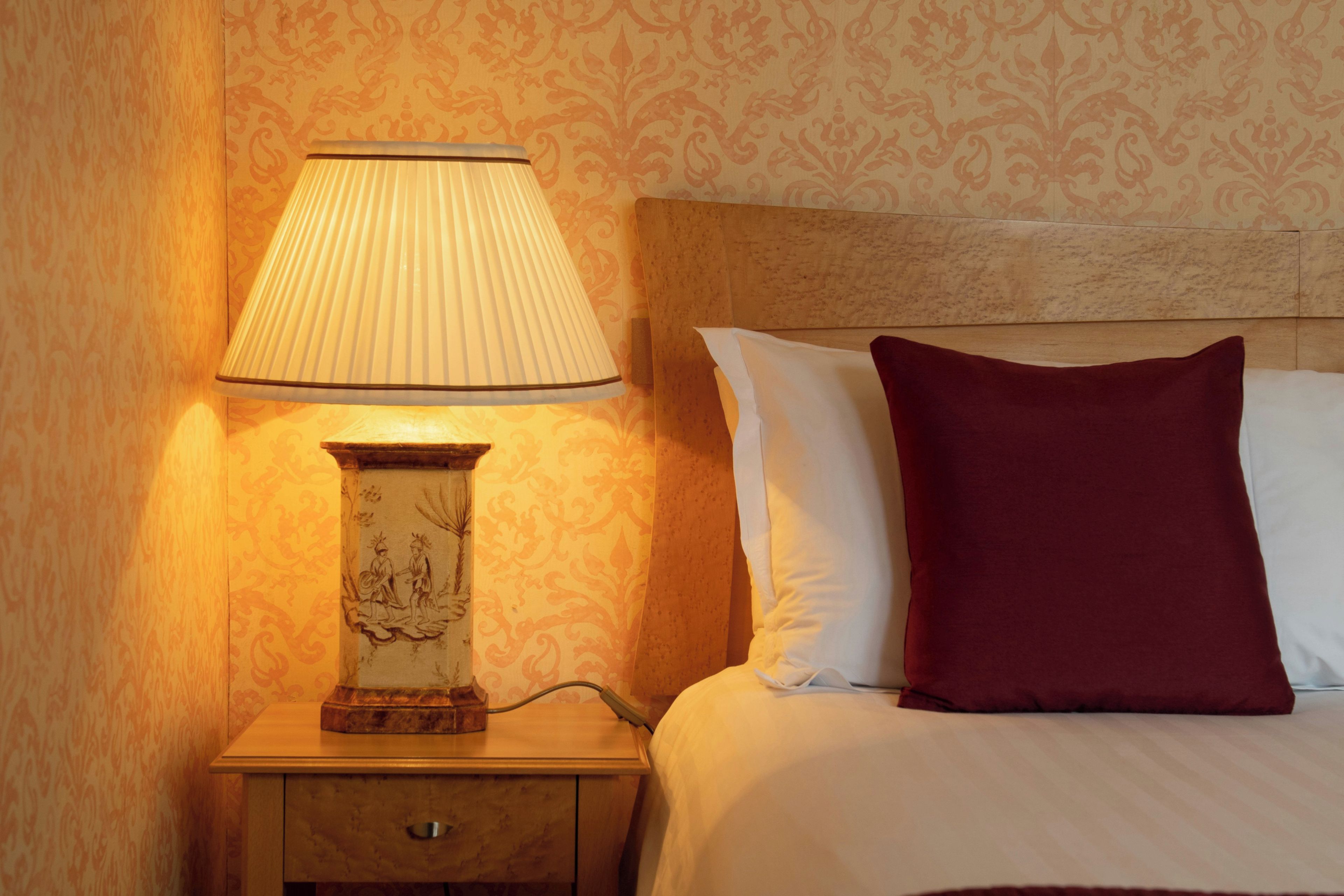 Habitación de hotel con lámpara de noche, mesita de noche de madera, cama con almohadas blancas y burdeos, y papel tapiz estampado.