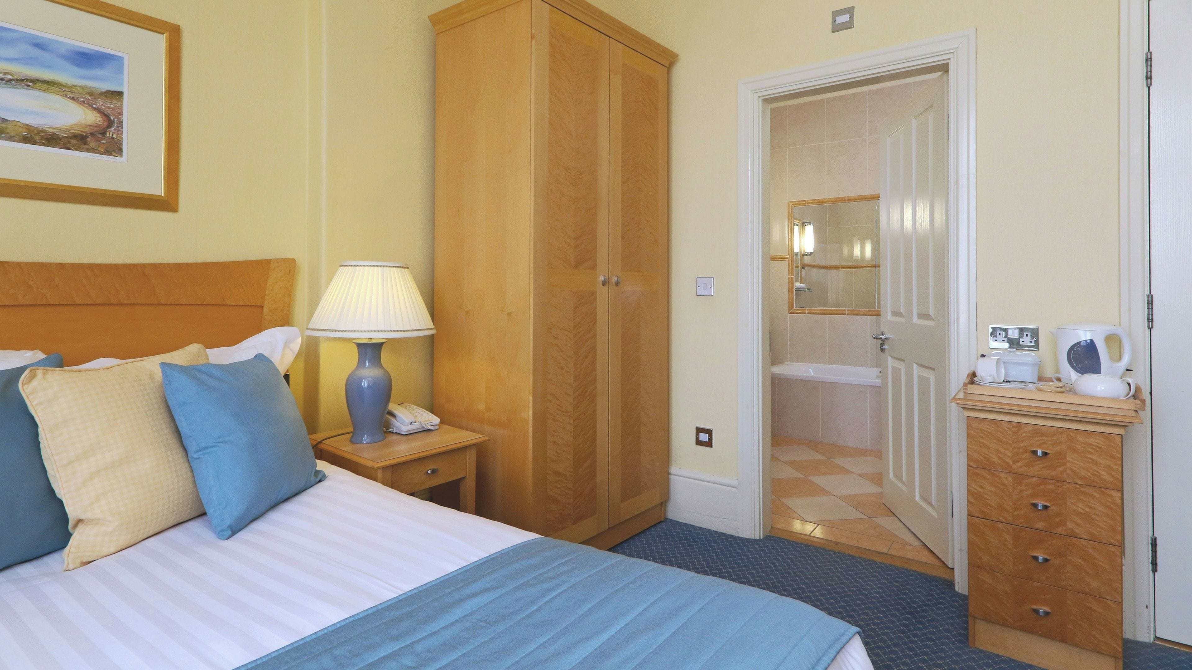 A hotel room with a bed, a nightstand with a lamp, a wardrobe, a bathroom with a mirror, and a small wooden dresser with a kettle.
