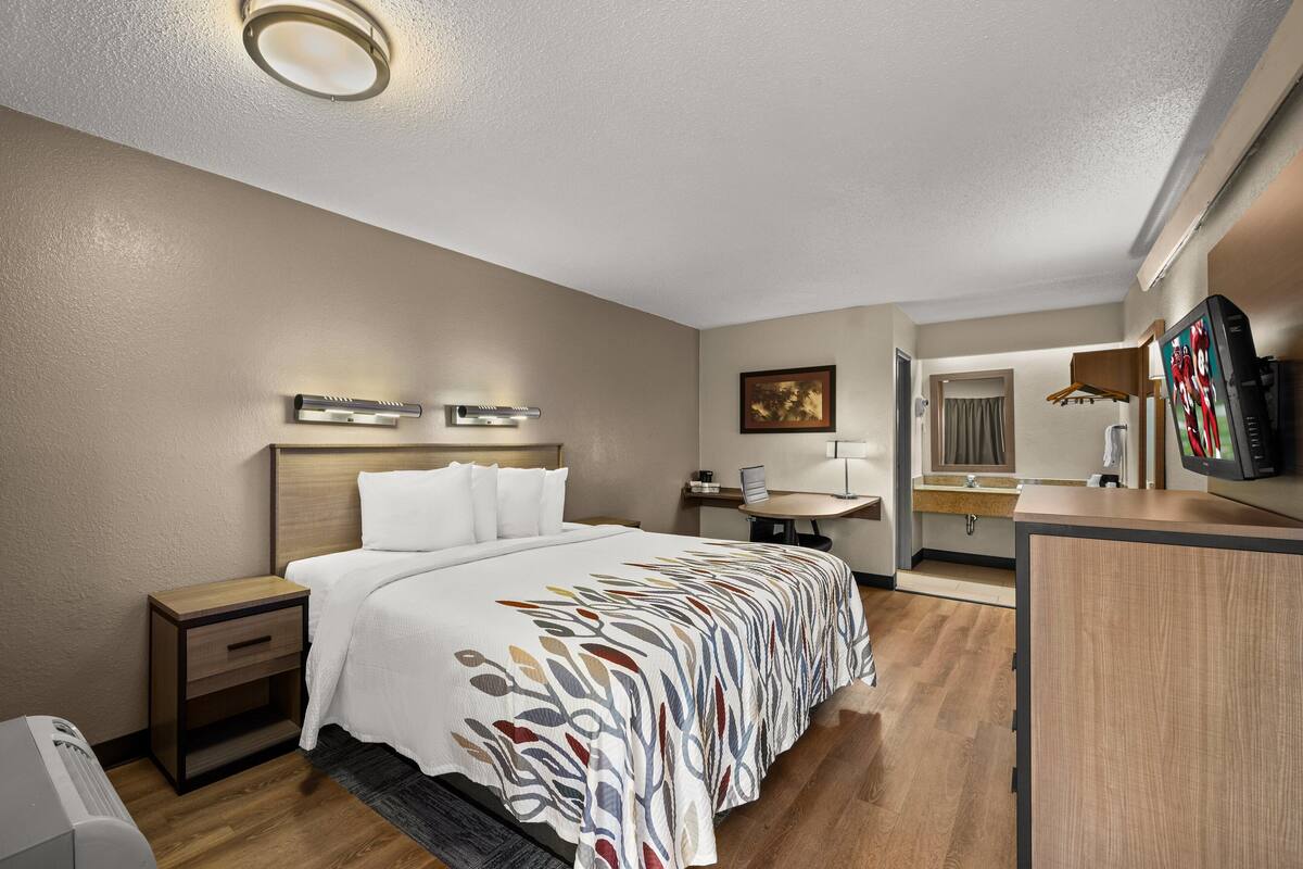 A hotel room with a bed, bedside table, lamp, mirror, and a desk with a chair.