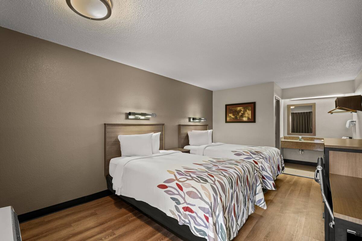 A hotel room with two beds, a wooden floor, a desk with a mirror, and a painting on the wall.
