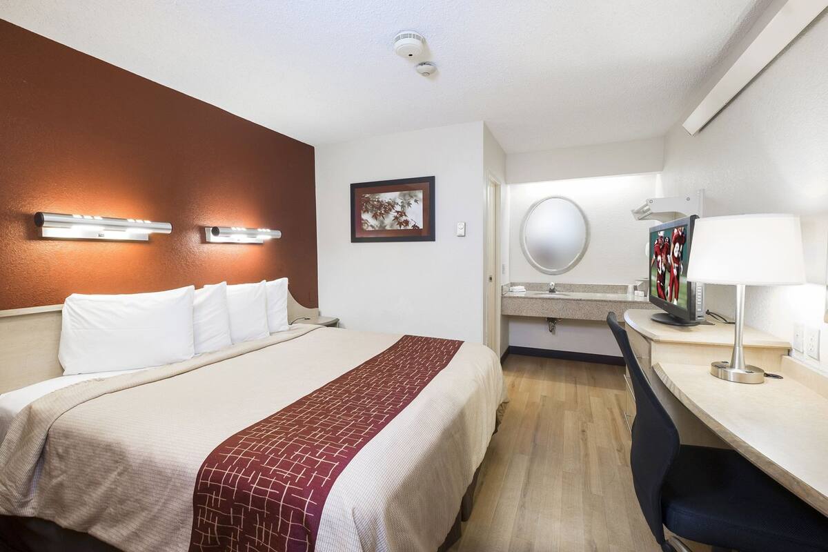 A hotel room with a bed, bedside lamps, a desk with a chair, a round mirror, and a television.